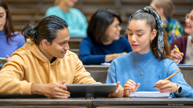 Foto von zwei Studierenden in einem Hörsaal, die miteinander sprechen und gemeinsam auf ein Tablet schauen. Im Hintergrund weitere Studierende.
