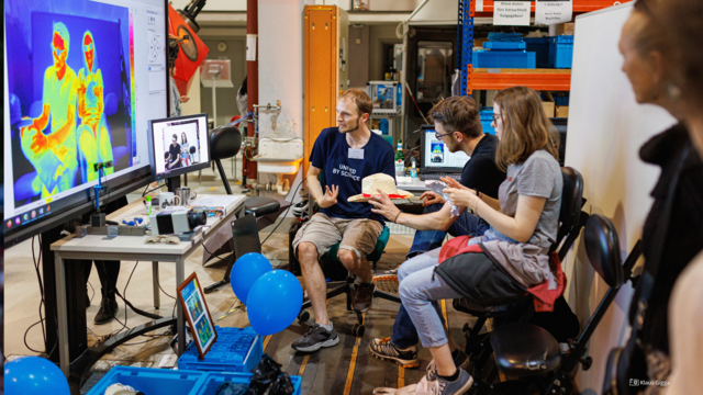 Foto von Menschen, die in einer Versuchshalle vor einem Bildschirm sitzen, auf dem sie zu sehen sind, wie sie von einer Wärmebildkamera aufgezeichnet werden