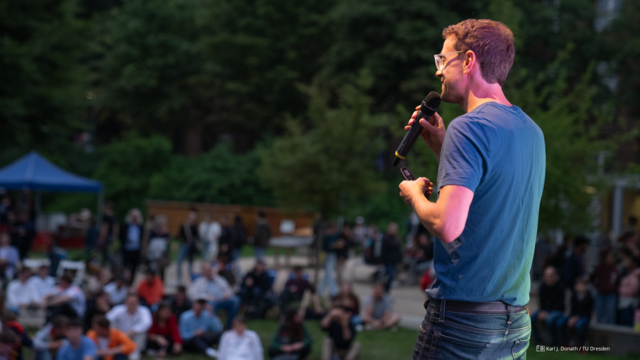 Foto von einem Mann auf einer Bühne mit Mikrofon in der Hand; im Hintergrund Publikum