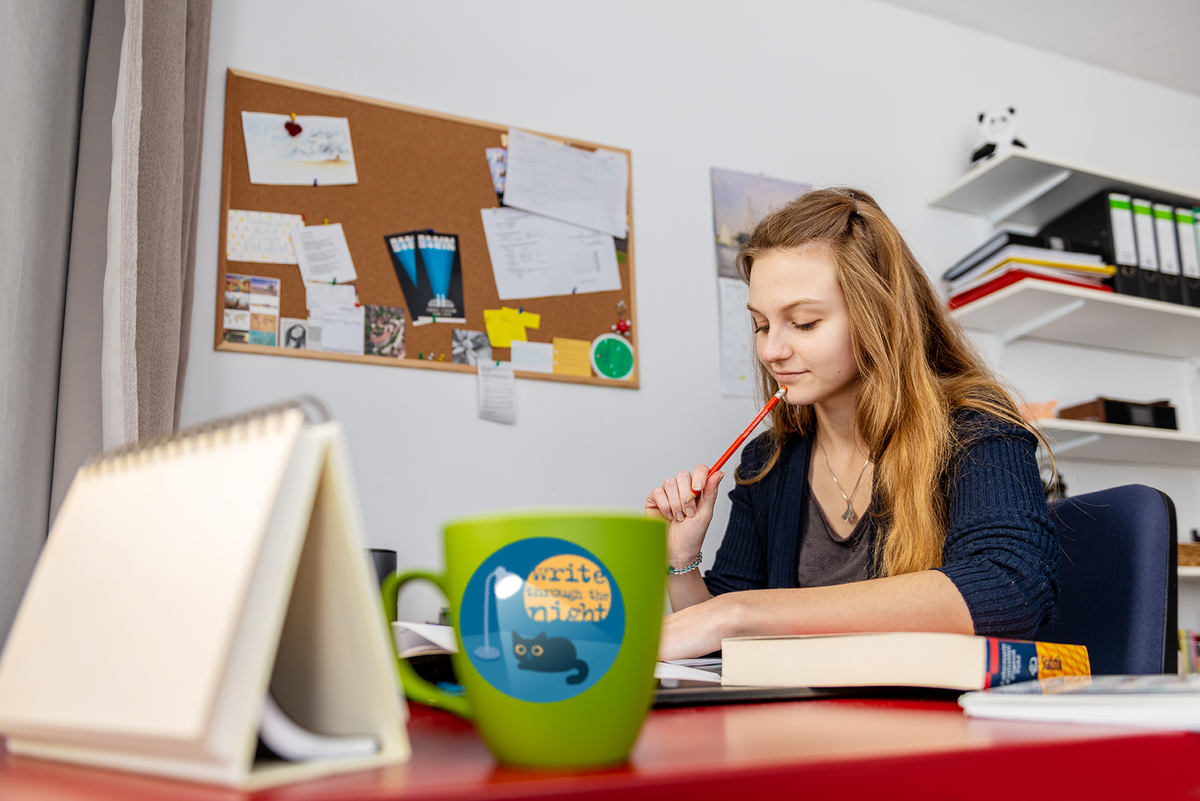 Eine junge Frau sitzt an am Schreibtisch. Sie hält einen Bleistift an ihren Mund. Sie blickt nachdenklich auf ihr Buch. Im Vordergrund sieht man eine Tasse, auf der die Grafik der Langen nacht des Schreibens zu sehen ist. Auf dieser sieht man eine Katze unter einer Lampe. Hinter ihr steht "Write through the night". Eine junge Frau sitzt an am Schreibtisch. Sie hält einen Bleistift an ihren Mund. Sie blickt nachdenklich auf ihr Buch. Im Vordergrund sieht man eine Tasse, auf der die Grafik der Langen nacht des Schreibens zu sehen ist. Auf dieser sieht man eine Katze unter einer Lampe. Hinter ihr steht "Write through the night".