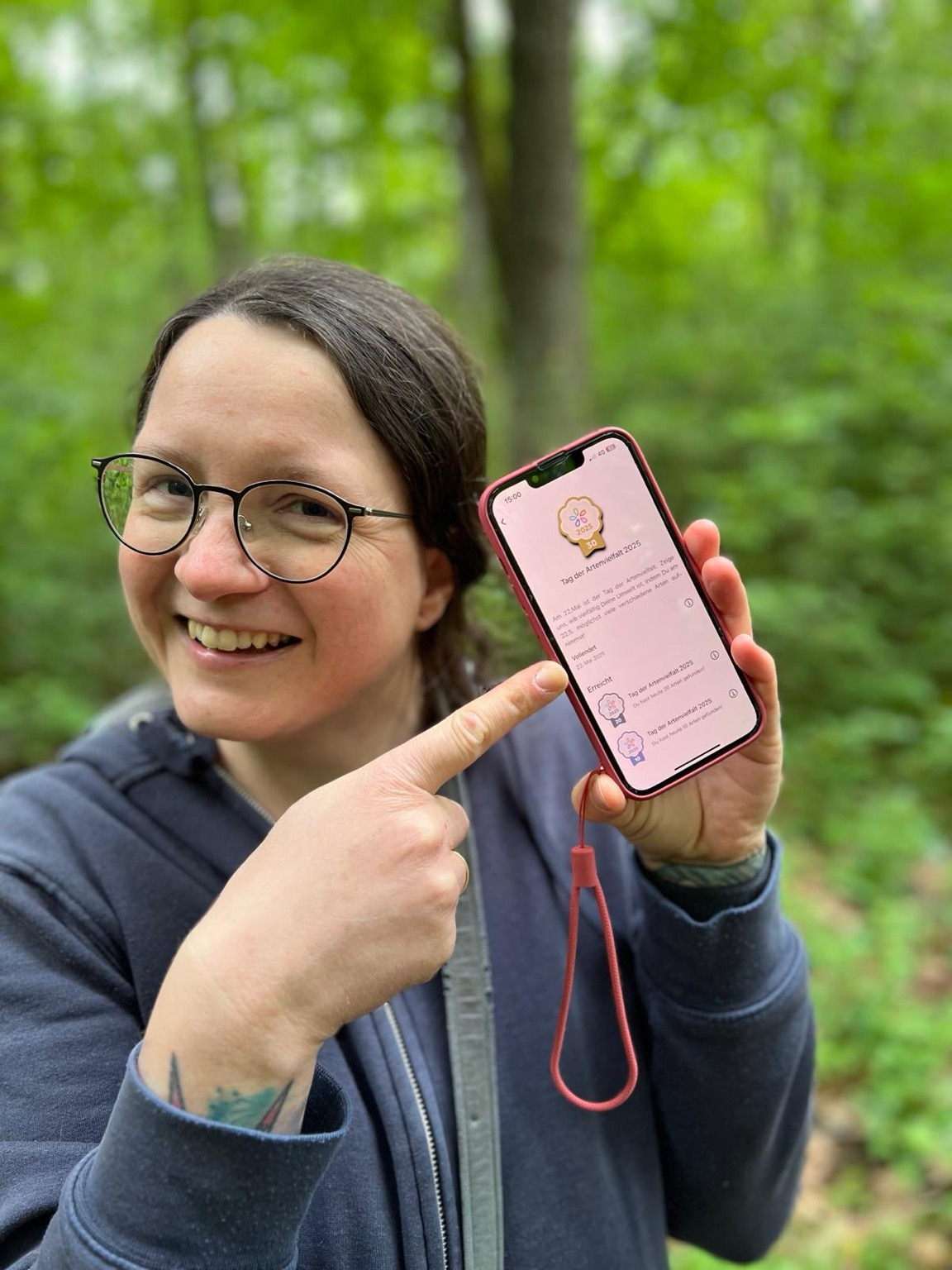 Eine lächelnde Frau mit blauem Kapuzenpulli und Brille zeigt auf ein Telefon, welches das Abzeichen präsentiert. Eine lächelnde Frau mit blauem Kapuzenpulli und Brille zeigt auf ein Telefon, welches das Abzeichen präsentiert.
