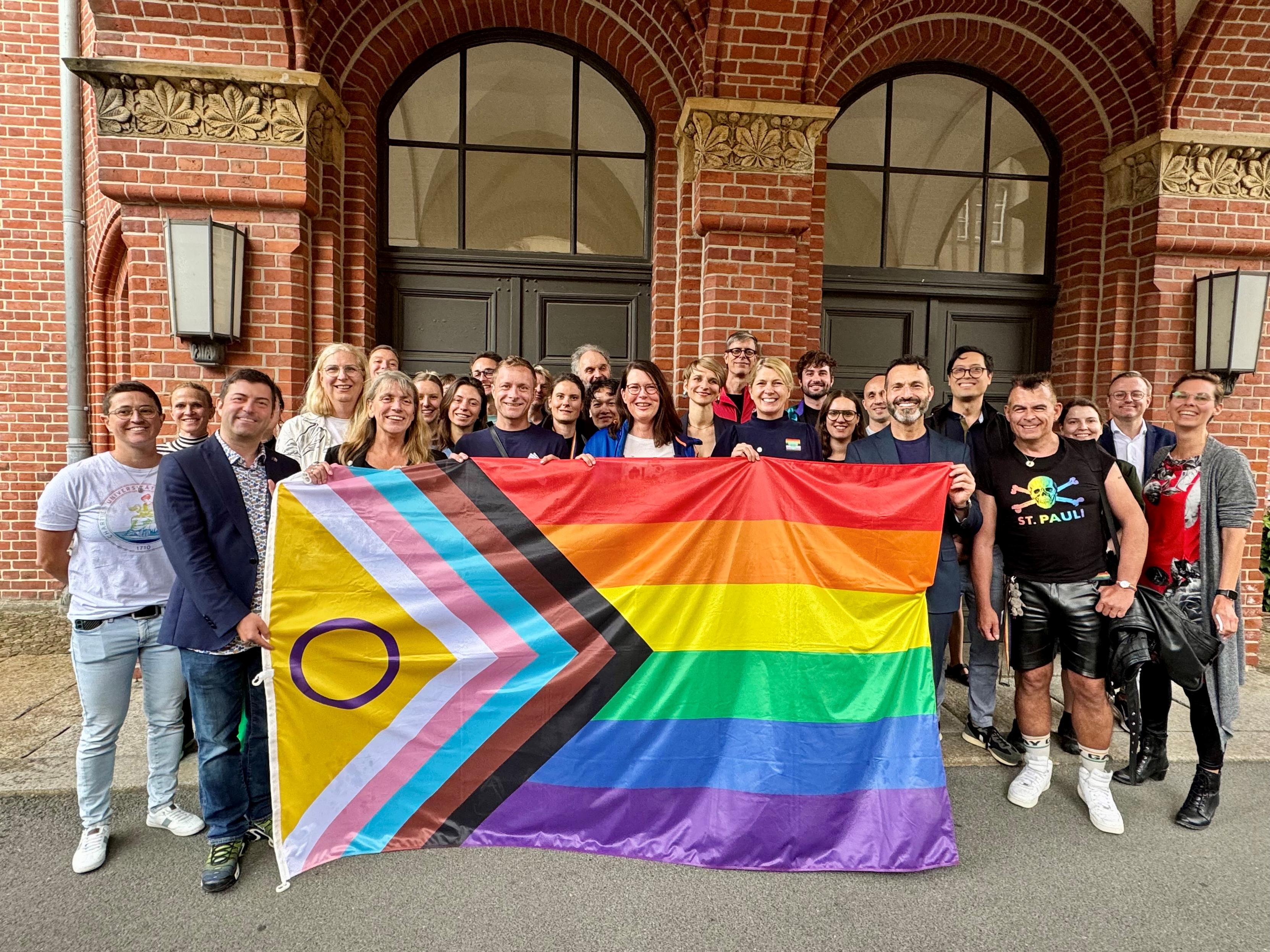 Eine Gruppe von Menschen steht vor einem Haus und hält die progressive Pride-Flagge in die Kamera. Die Personen lächeln.