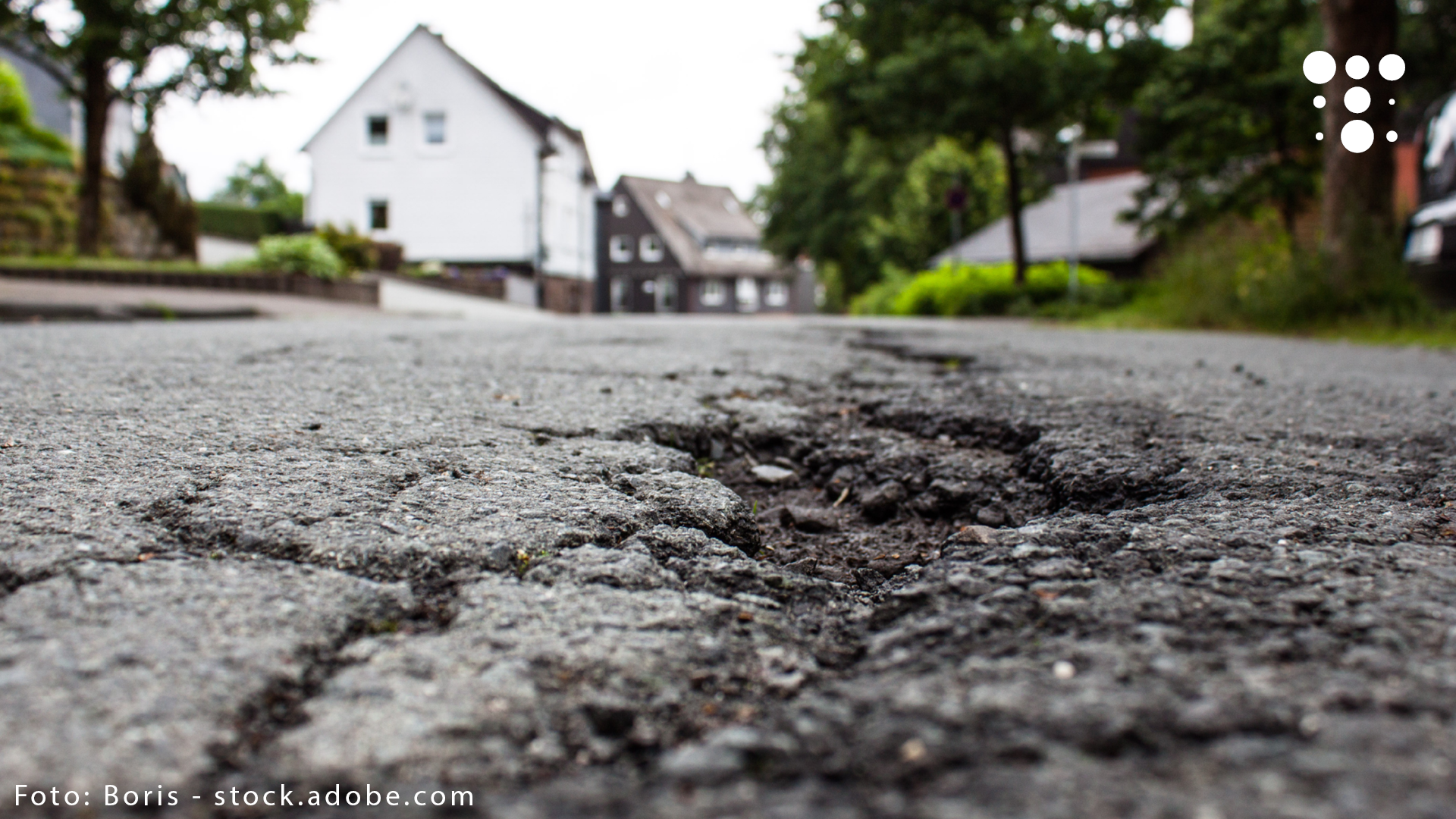 Nahaufnahme einer Asphaltstraße mit Schlaglöchern und unscharfem Hintergrund mit Bäumen und Wohnhäusern