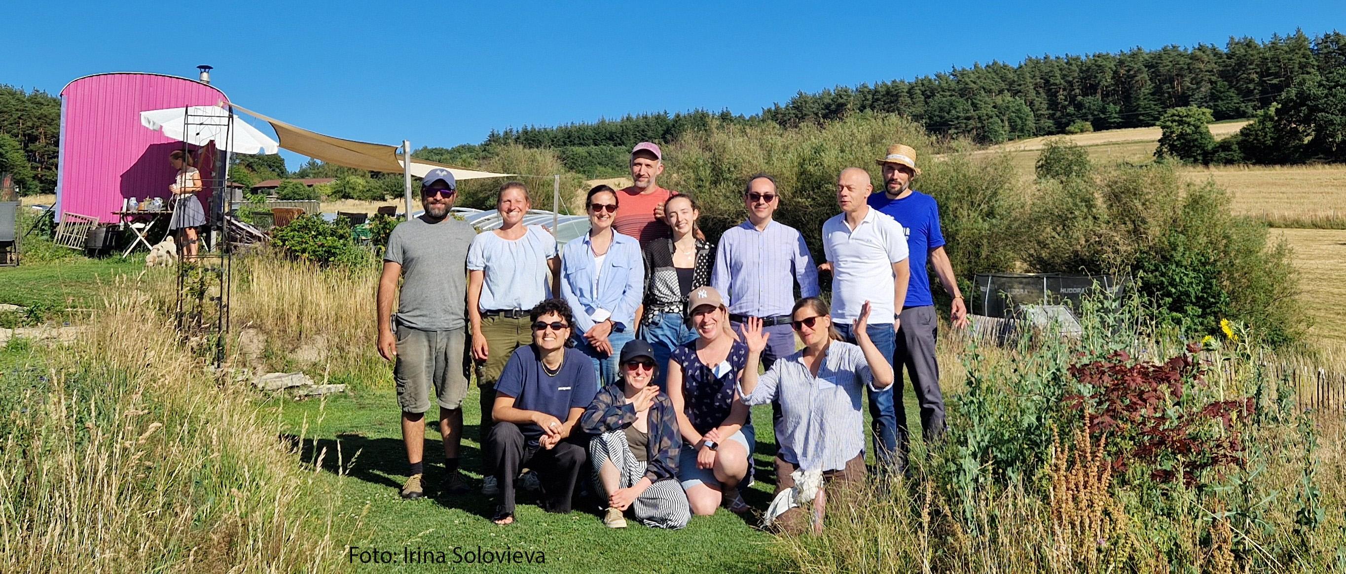 Eine Gruppe von elf Personen posiert bei Sonnenschein auf einer Wiese vor einer ländlichen Kulisse mit einem pinken Wohnwagen.